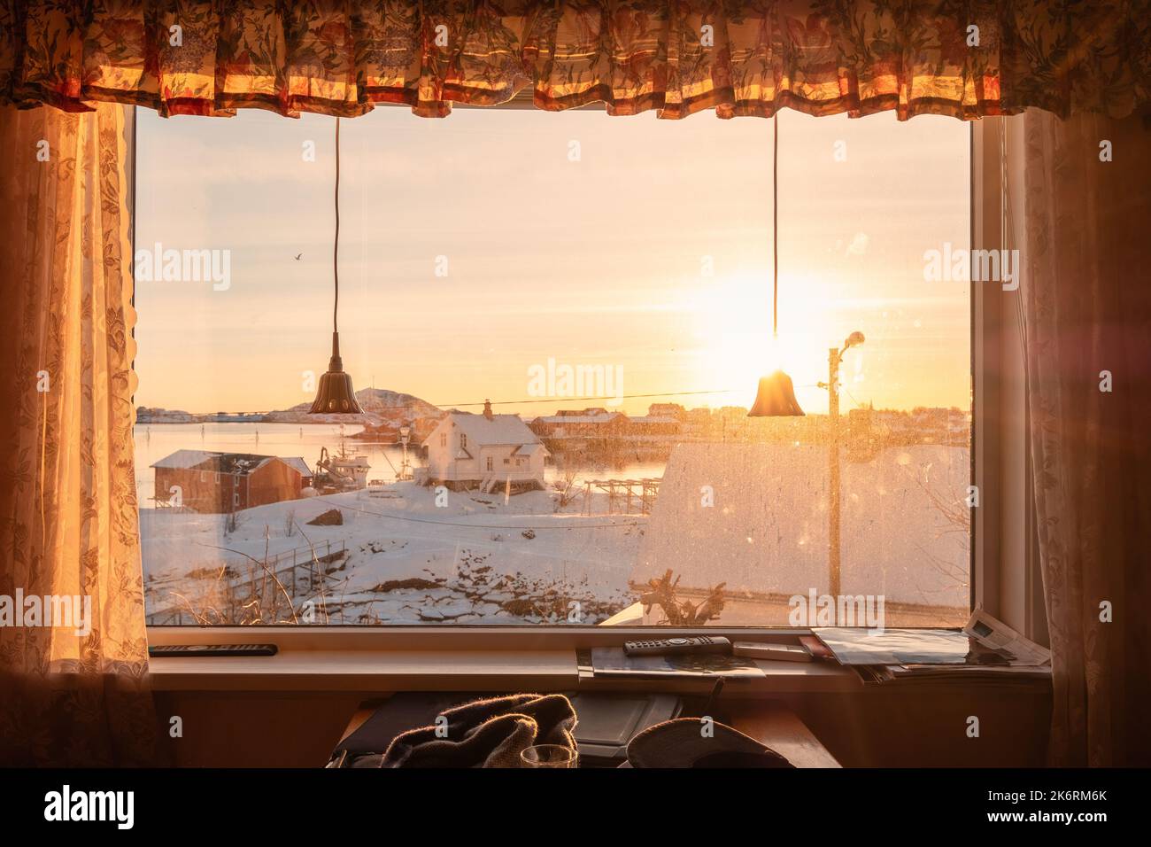 Blick vom Vorhangfenster mit Sonnenuntergang über dem verschneiten Dorf an der Küste in der Wintersaison. Nostalgie und Heimweh Stockfoto