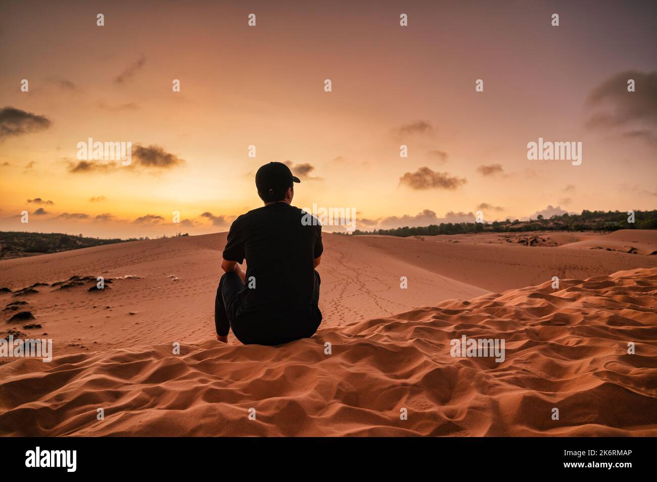 Rückansicht eines jungen asiatischen Mannes mit Hut, der während des Sonnenuntergangs in Muine, Vietnam, auf einer Sanddüne sitzt Stockfoto