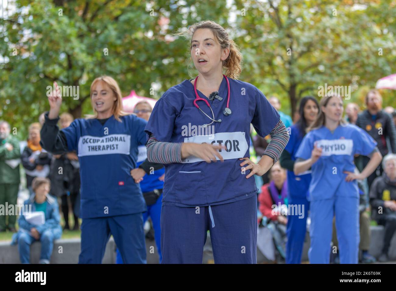 London, Großbritannien. Oktober 2022. Eine Gruppe medizinischer Fachkräfte im Vereinigten Königreich, darunter Ärzte und ein Physiotherapeut, nimmt an einer öffentlichen Demonstration Teil. Sie tragen gekennzeichnete Uniformen und Stethoskope, um ihre Rolle visuell darzustellen. Die Veranstaltung findet unter freiem Himmel im Herbstlaub statt, mit einer Menge von Fans im Hintergrund. Ärzte und Wissenschaftler für die Extinction Rebellion inszenieren A sterben in der Nähe des London Eye auf der Southbank. Penelope Barritt/Alamy Live News Stockfoto