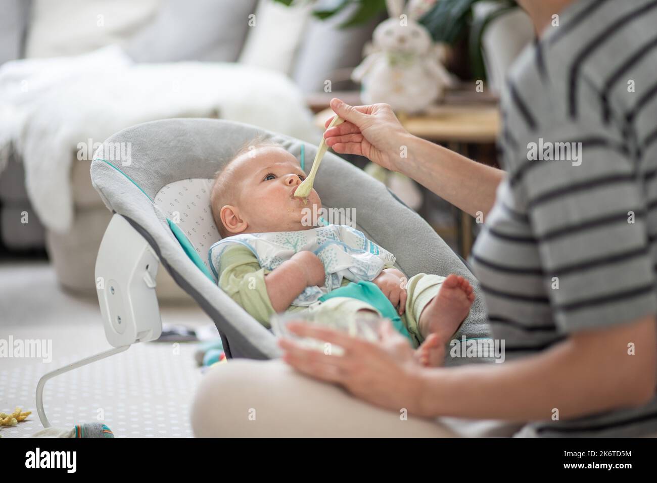 Mutter Löffel Fütterung ihres kleinen Jungen Kleinkind in Babystuhl mit Fruchtpüree. Baby feste Nahrung Einführung Konzept. Stockfoto