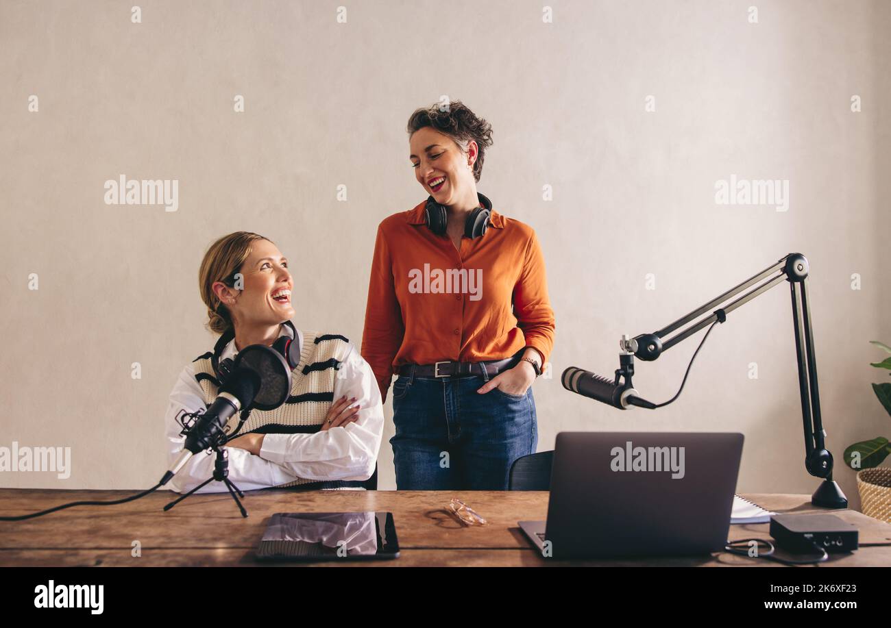 Fröhliche Podcasters, die sich gegenseitig mit Headsets um den Hals lächeln. Fröhliche Frauen, die eine Audioübertragung in einem Heimstudio veranstalten. Zwei weibliche Kontra Stockfoto