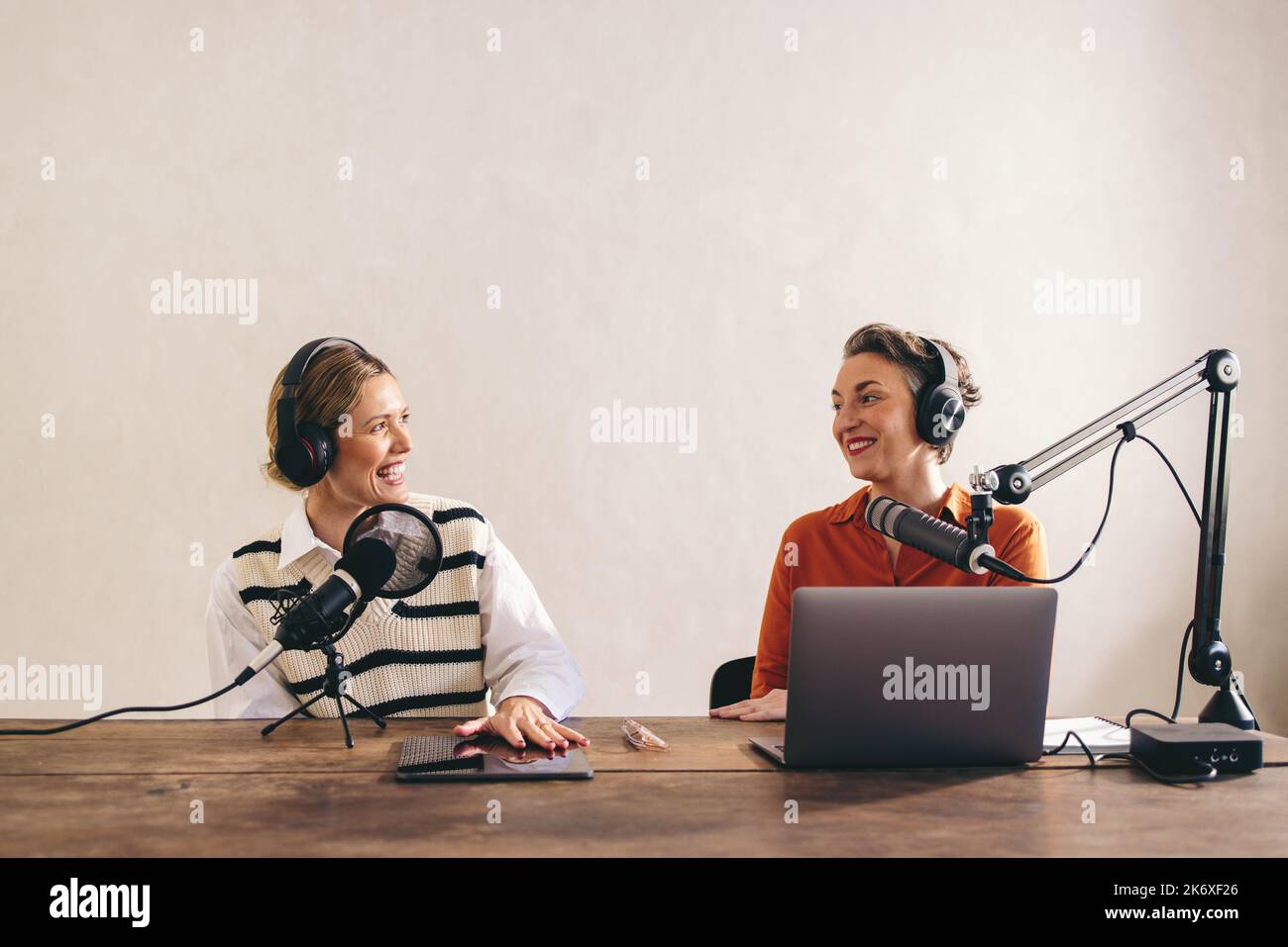 Zwei weibliche Content-Macher lächeln fröhlich, während sie einen Internet-Podcast in einem Heimstudio aufnehmen. Zwei fröhliche Frauen, die gemeinsam eine Audioübertragung für moderiert haben Stockfoto