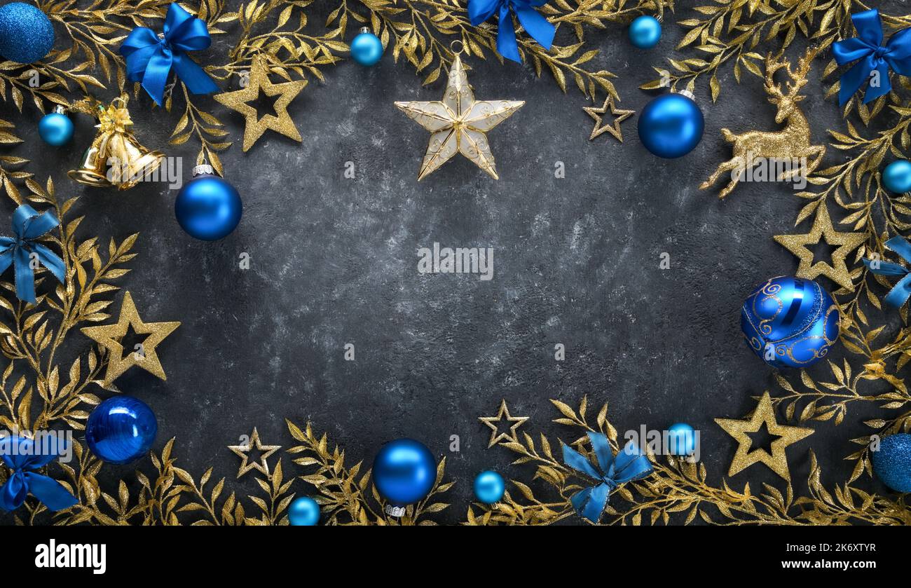 Weihnachtshintergrund mit eleganten Goldverzierungen als Rahmen, blauen Kugeln und Schleifen, auf einem dunkelgrauen strukturierten Brett als Kopierraum Stockfoto