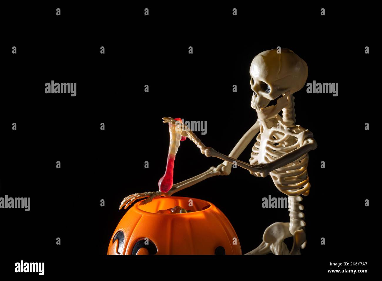 Ein sitzendes Skelett zieht sich aus einem Eimer in Form eines Kürbisbonbons auf schwarzem Hintergrund, Kopierraum. Halloween Trick or Treat Stockfoto