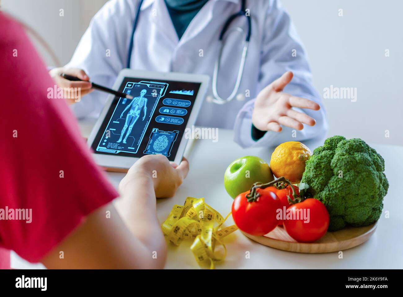 Ernährungsdozentin mit digitalem mobilen Tablet mit virtuellem grafischem Symboldiagramm und Gemüse und Obst mit Patient am Schreibtisch im Krankenhaus Stockfoto
