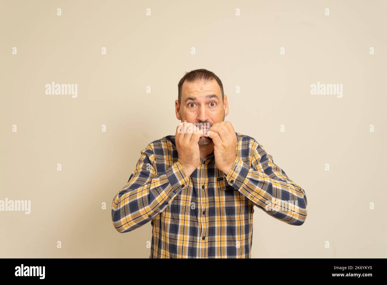 Studioportrait eines verängstigten und schockierten Mannes in seinem 40s-er, der seine Nägel beißt, mit weiten Augen auf etwas Schreckliches starrt, in einem legeren Hemd, das über Beige steht Stockfoto