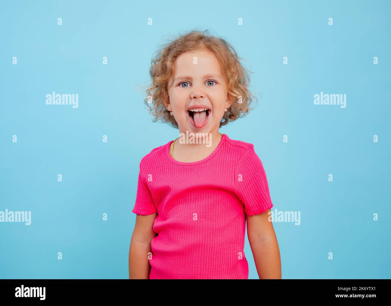 Kleines, wenig expressives, verspieltes, positiv lockiges blondes Mädchen im rosa Hemd ragt aus der Zunge und grimazing zur Kamera im Studio Stockfoto