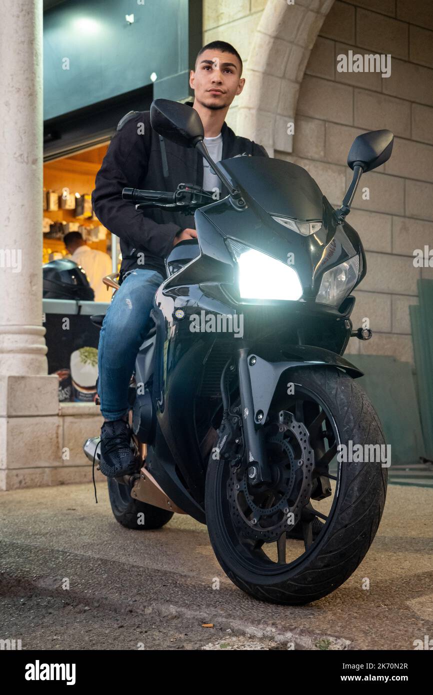 Ramallah, Ramallah und al-Bireh Governorate, Palästina, 12. Juli 2022: Junger, hübscher arabischer Mann in blauer Jeans und schwarzer Jacke sitzt auf einem schwarzen Motorcycl Stockfoto