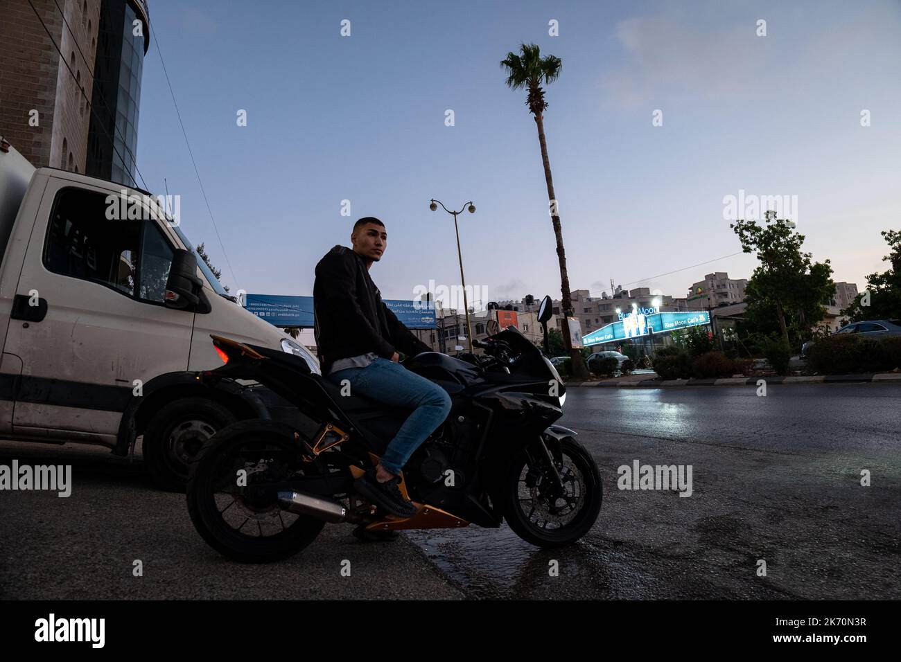 Ramallah, Ramallah und al-Bireh Governorate, Palästina, 12. Juli 2022: Junger, hübscher arabischer Mann in blauer Jeans und schwarzer Jacke sitzt auf einem schwarzen Motorcycl Stockfoto