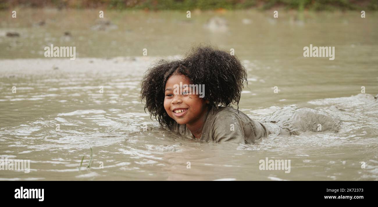 Fröhliches afroamerikanisches Kindermädchen, das während der Regenzeit in einer feuchten Schlammpfütze spielt. Stockfoto