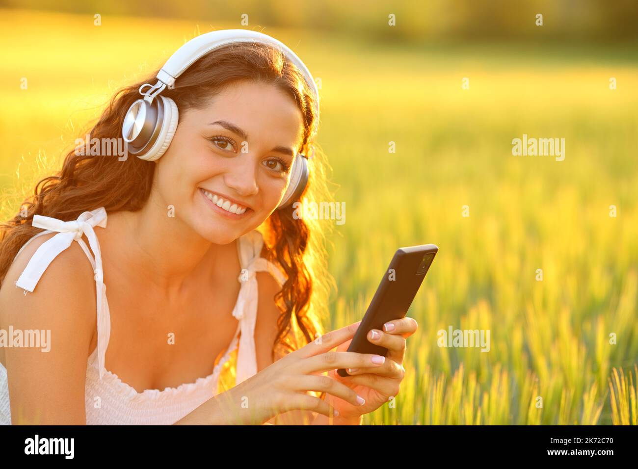 Eine glückliche Frau, die Kopfhörer trägt und Musik hört, sieht Sie auf einem Feld an Stockfoto