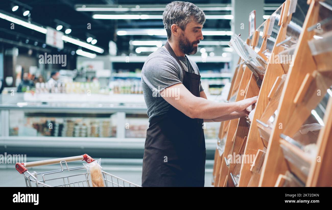 Ein vielbeschäftigter männlicher Verkäufer legt in der Bäckerei im Lebensmittelgeschäft Brot in die Regale, ein bärtiger Mann trägt eine Schürze. Verkauf von Produkten, Beruf und Handelskonzept. Stockfoto