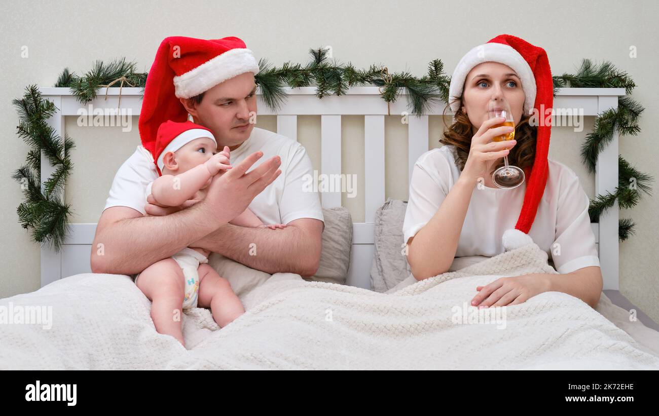 Frau trinkt Alkohol, während ein Mann mit einem Kind in einem Bett sitzt, das zu Weihnachten und Neujahr dekoriert ist, Familienpaar in einem Schlafzimmer Stockfoto