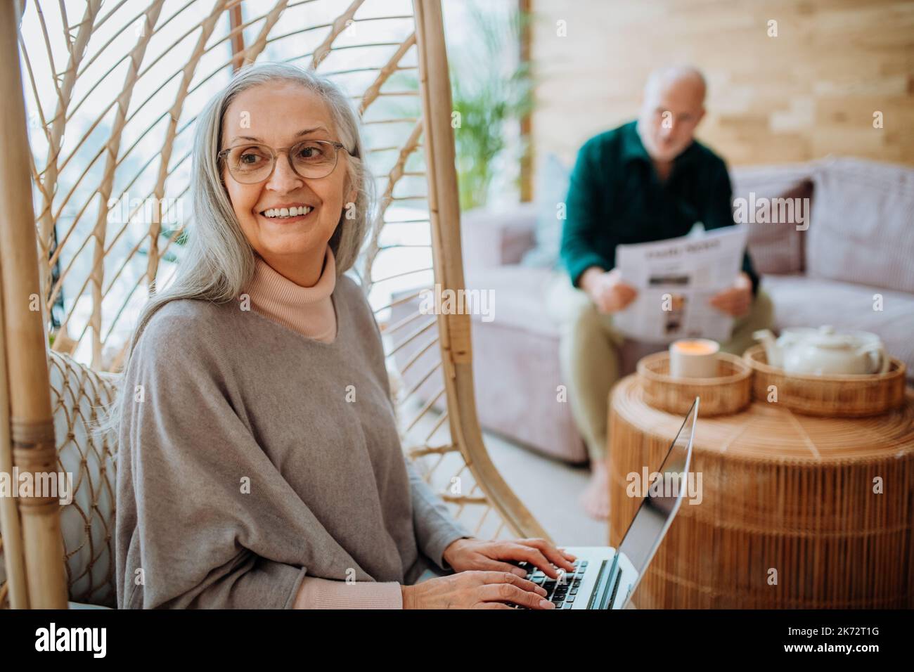 Ältere Frau, die in einer Schaukel im Freien sitzt und am Laptop arbeitet, ihr Mann ruht sich auf dem Sofa aus. Konzept des Heimbüros und eines aktiven Senior-Lebensstils. Stockfoto