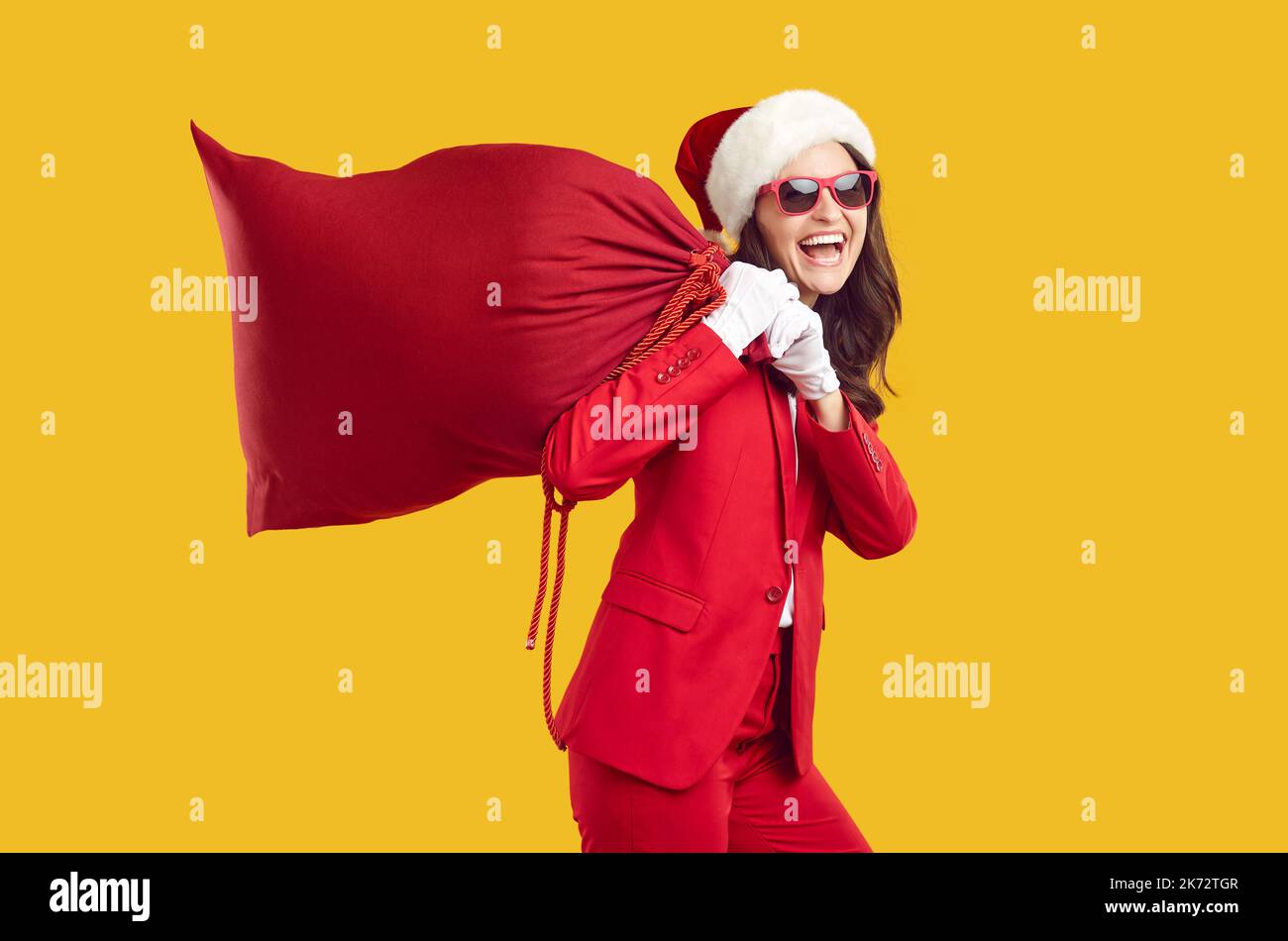 Lustige moderne Weihnachtsfrau, die laut lacht und eine große rote Tasche mit Geschenken auf ihren Schultern trägt. Stockfoto
