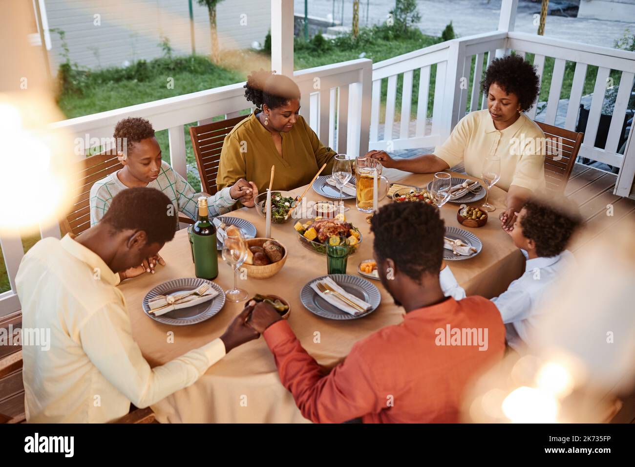 Blick aus der Perspektive der afroamerikanischen Familie, die beim Abendessen im Freien Anmut sagte und die Hände in gemütlicher Abendumgebung hielt Stockfoto