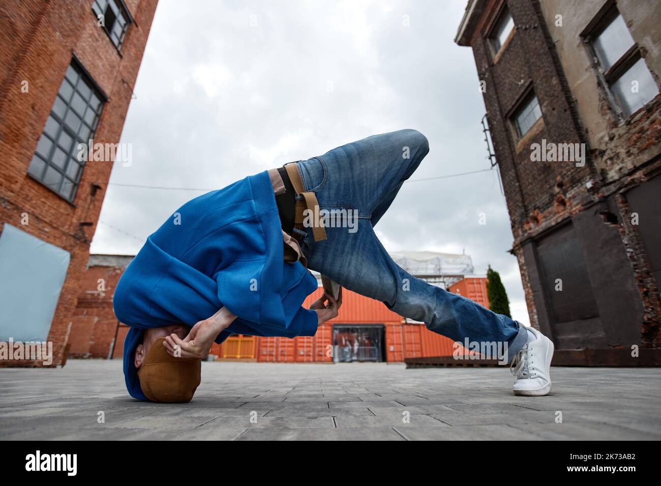 Ganzkörperaufnahme eines jungen Mannes, der in einer urbanen Fabrik im Freien Breakdance-Pose macht und sein Gesicht versteckt Stockfoto