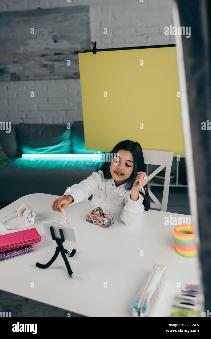 Brunette Mädchen hält Perlen Armband in der Nähe von Smartphone auf Halter und Bleistifttaschen mit kabellosen Kopfhörern Stockfoto