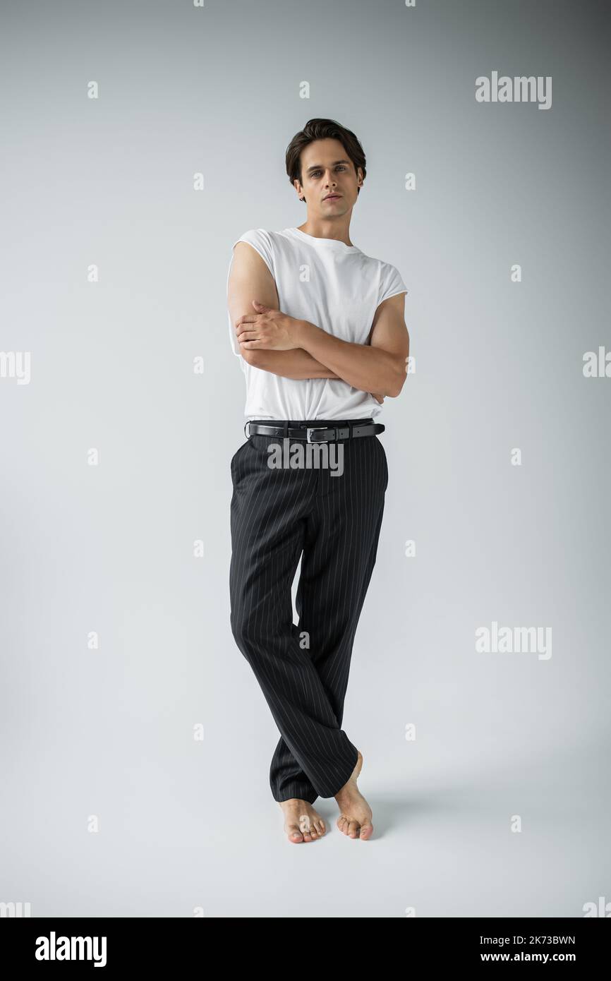 Barfuß-Mann in durchgehender Länge in gestreifter Hose und weißem T-Shirt, das mit gekreuzten Armen auf grau posiert Stockfoto