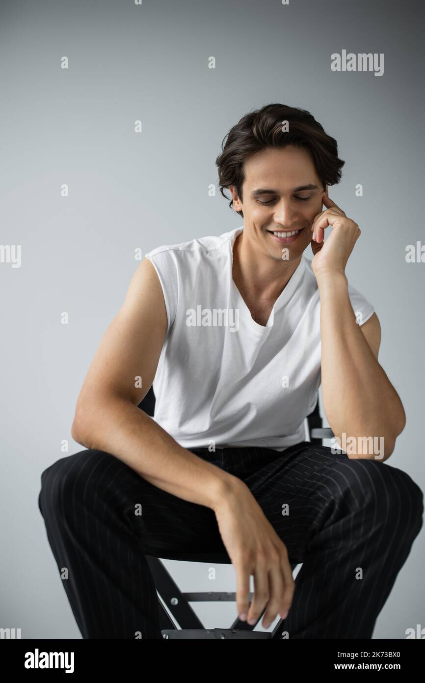 Fröhlicher Mann in gestreifter Hose und weißem T-Shirt auf dem Lagerstuhl in Grau Stockfoto