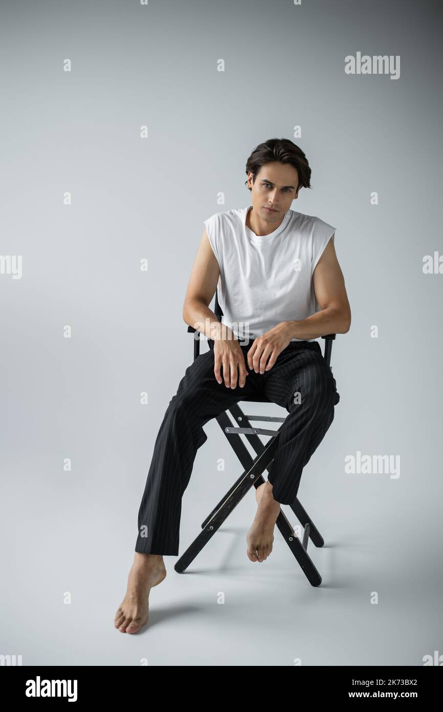 In voller Länge barfuß Mann in Hosen und weißem T-Shirt auf Lagerstuhl auf grau sitzen Stockfoto