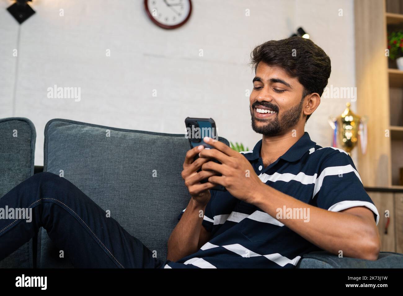 Glücklich lächelnder junger Mann, der Mobiltelefon benutzt, während er zu Hause auf dem Sofa sitzt - Konzept von Social Media Sharing, Internet und Entspannung Stockfoto