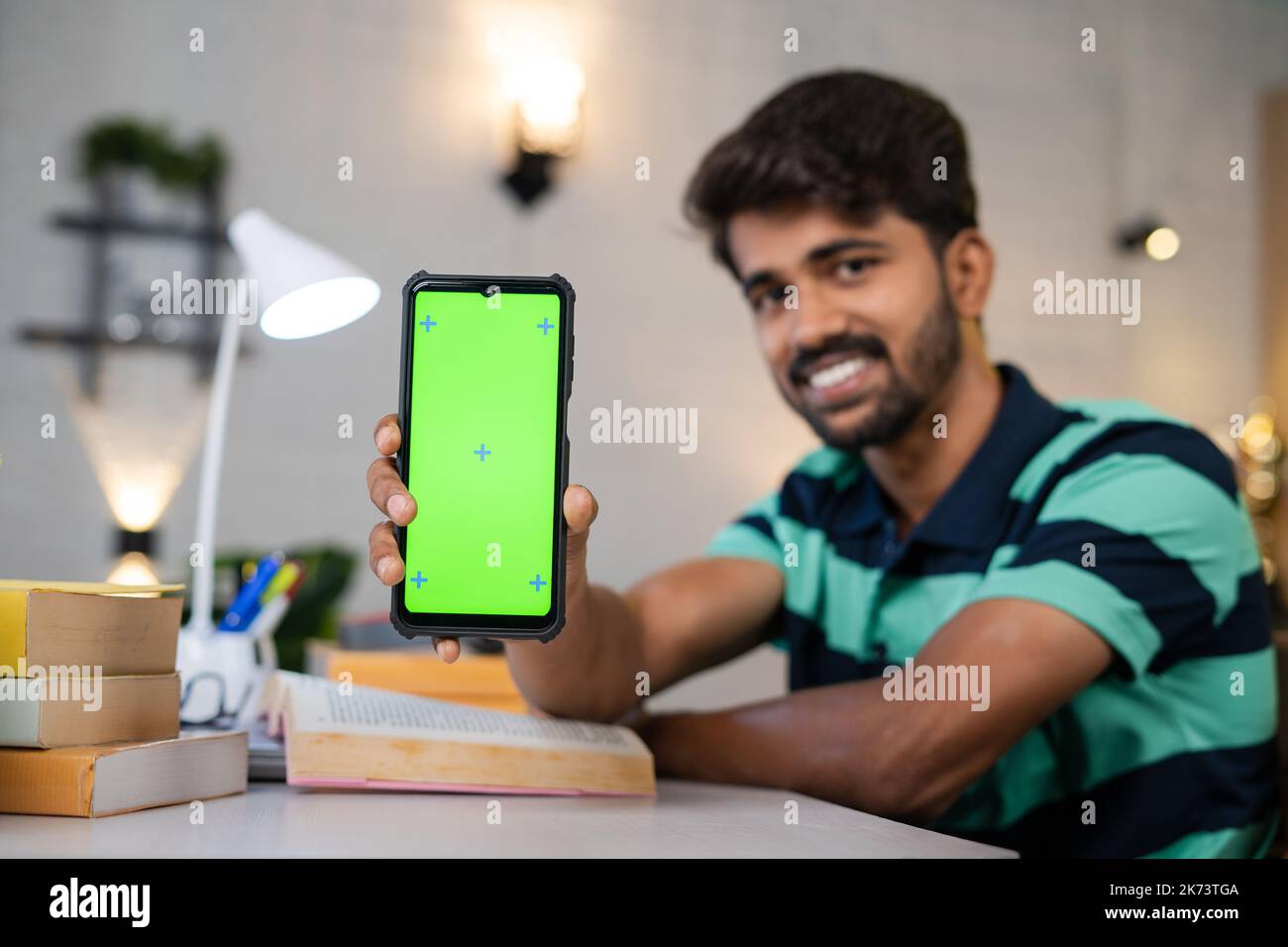 Glücklich lächelnder Student am Lerntisch mit Büchern zeigen Greenscreen-Handy durch Blick auf Kamera - Konzept der App-Förderung, Online-Buchung und Stockfoto