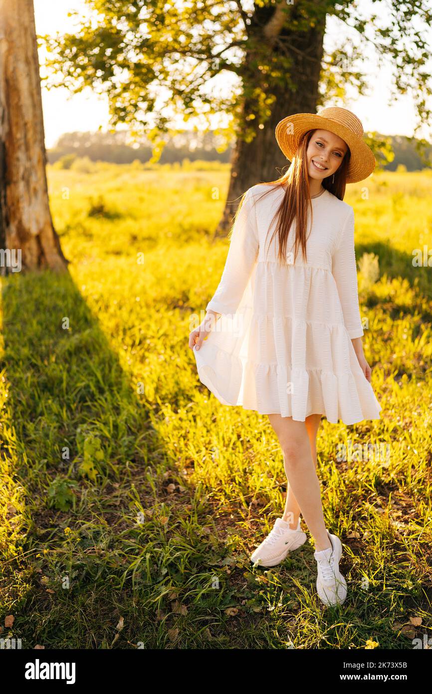 Vertikales Porträt einer romantischen jungen Ingwer-Frau, die Strohhut und weißes Kleid trägt, das auf einer schönen Wiese mit grünem Gras steht und anschaut Stockfoto
