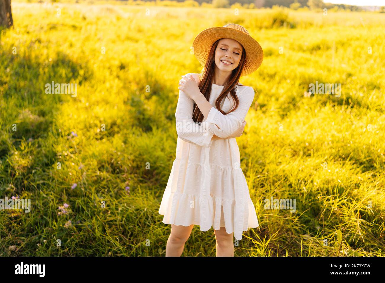 Porträt einer eleganten glücklichen Rothaarigen Frau in Strohhut und weißem Kleid, das mit geschlossenen Augen auf einer schönen Wiese aus grünem Gras posiert. Stockfoto