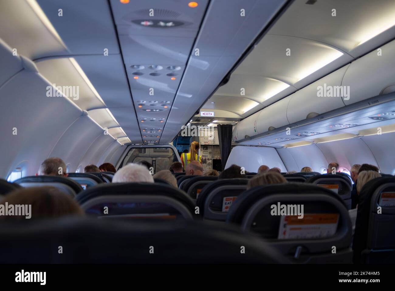 In der Passagierkabine eines easyJet Airbus A320 Airliner-Düsenflugzeugs mit Sitzen und Passagieren. Weibliches easyJet-Bodenpersonal, das sich mit der Crew begibt Stockfoto