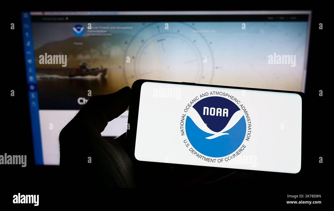 Person mit Mobiltelefon und Logo der National Oceanic and Atmospheric Administration (NOAA) auf dem Bildschirm vor der Webseite. Konzentrieren Sie sich auf die Telefonanzeige. Stockfoto