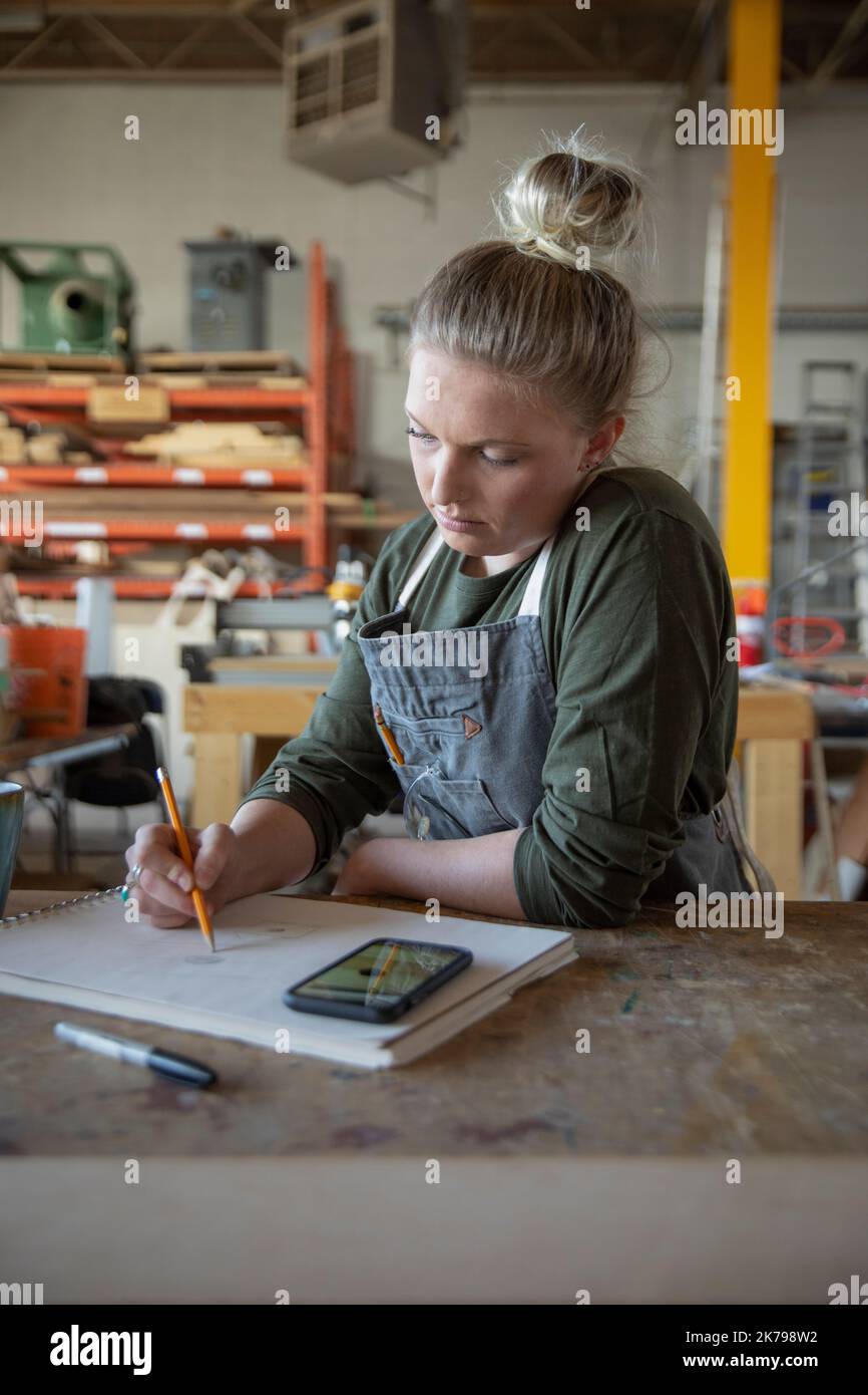 Junge Frau, die auf das Telefon schaut und in der Werkstatt Skizzenblock benutzt Stockfoto