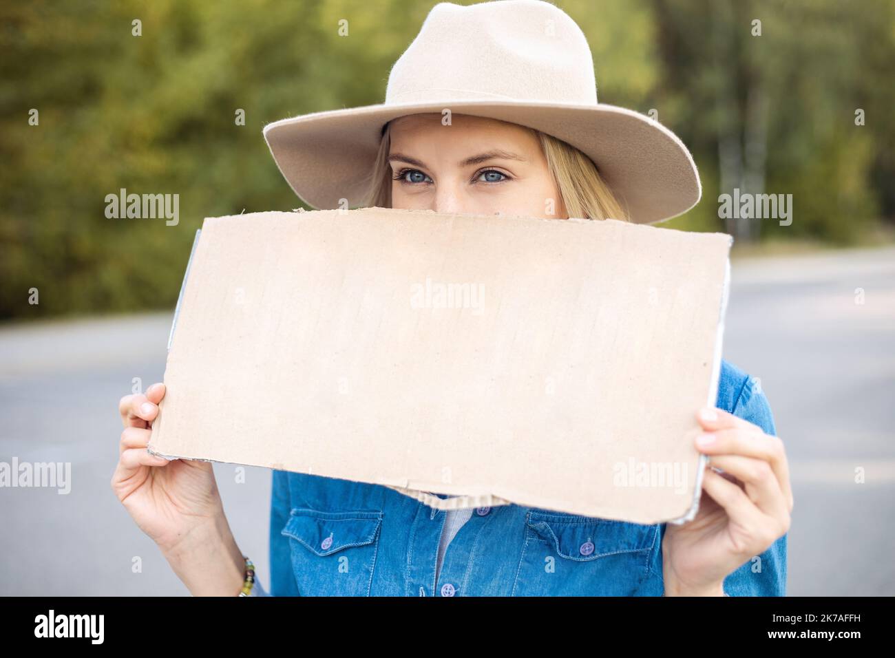 Porträt Nahaufnahme einer Frau, die hoffentlich auf das vorbeifahrende Auto im Wald wartet und leeres Pappplakat auf der Straße hält. Lady in Hut Flucht aus der Stadt durch Auto-Stop zu Stockfoto