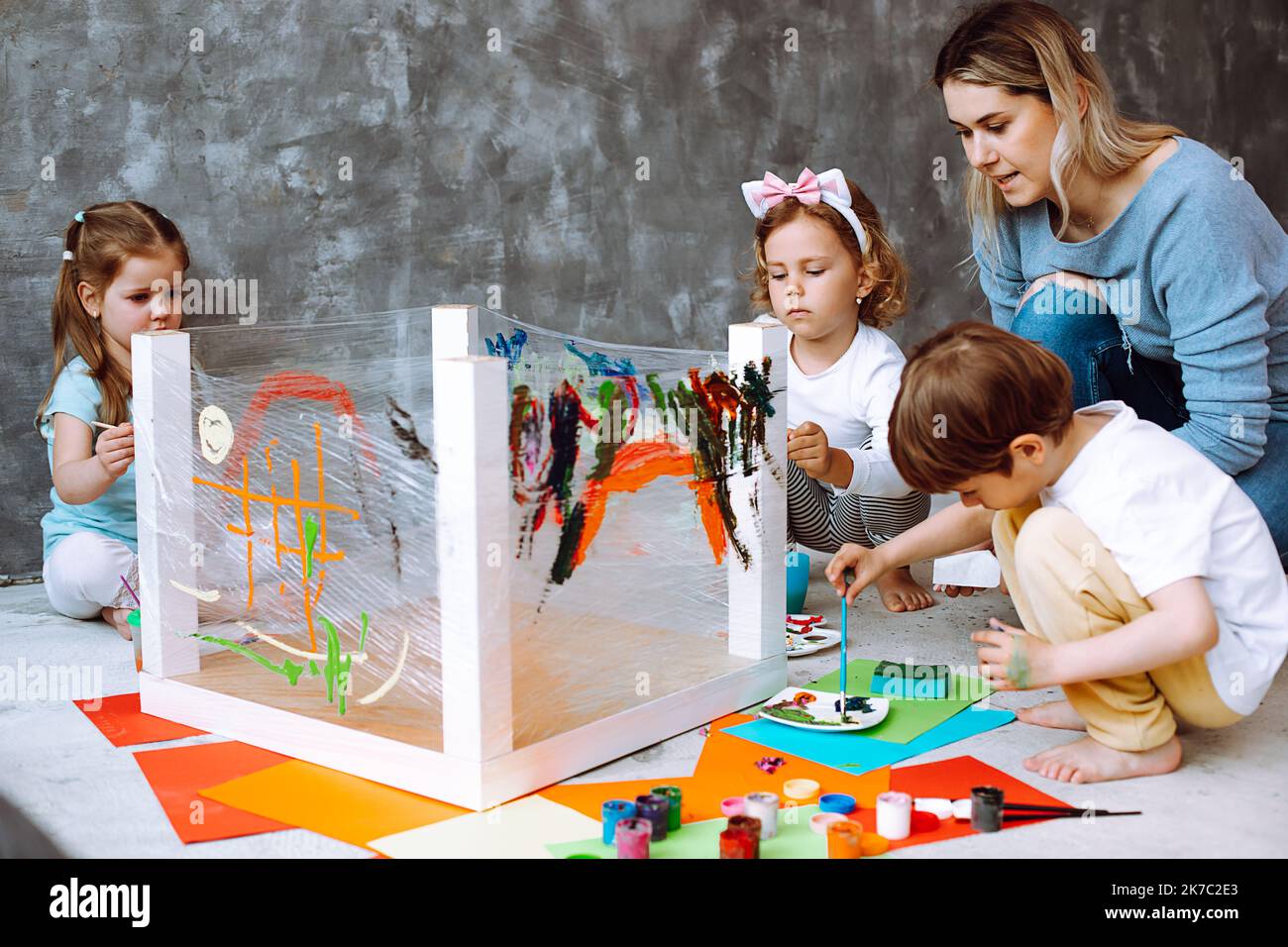 Erzieher, der Unterricht mit Kindern im Zeichnen im Kindergarten durchführt. Kinder sitzen im Spielzimmer und malen mit einem Pinsel auf einem Klebestreifen Stockfoto