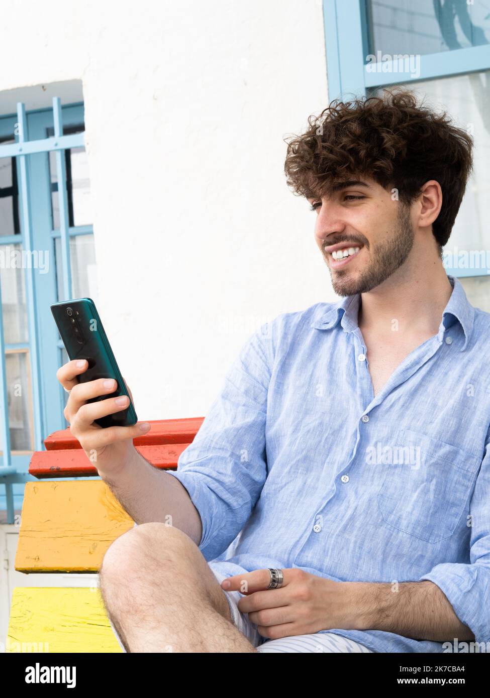 Lächelnder junger Mann, der auf einer Bank sitzt und mit einem Smartphone in einer Stadt chattet Stockfoto