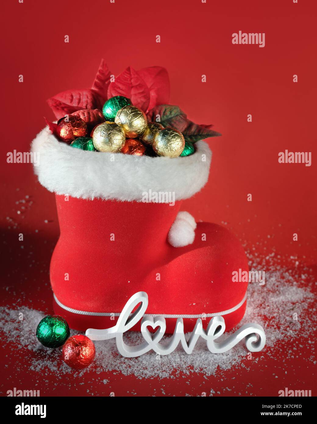 Roter Weihnachtsstiefel mit Schokoladen-Weihnachtskugeln. Holzwort Liebe auf rotem Hintergrund mit Schnee. Frohe Weihnachten Copy-Space, Platz für Text. Stockfoto