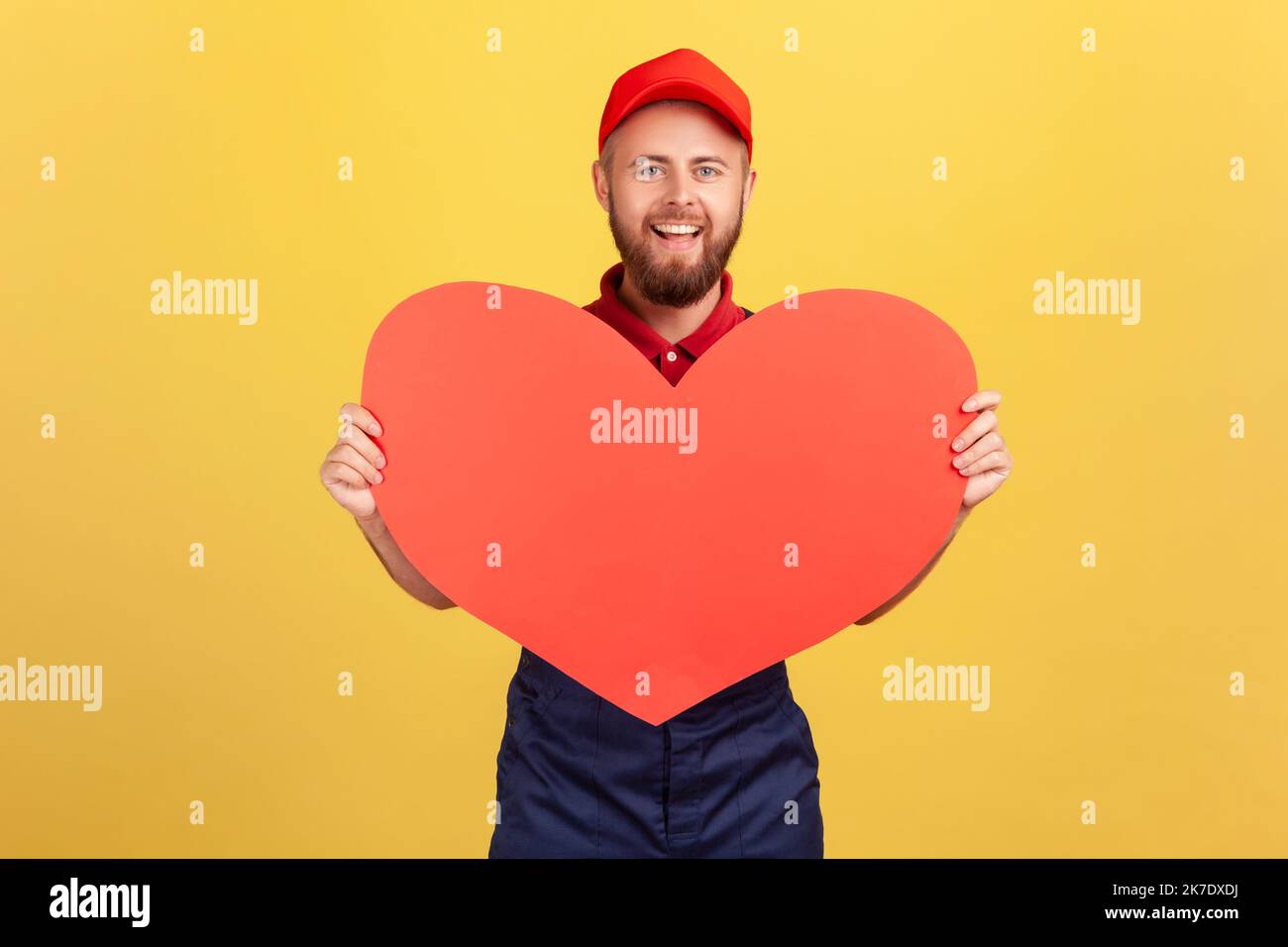 Porträt eines lächelnden romantischen Arbeiters mit blauen Overalls und roter Mütze mit großem roten Herzen, schnelle Geschenke während der Feiertage. Innenaufnahme des Studios isoliert auf gelbem Hintergrund. Stockfoto