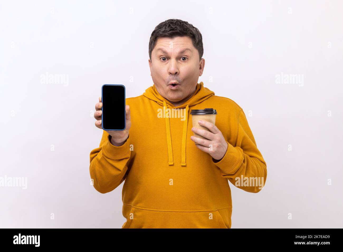 Porträt eines erstaunt überraschten Mannes mittleren Alters hält Smartphone mit leerem Bildschirm und Einweg-Tasse Kaffee, trägt urban Stil Hoodie. Innenaufnahme des Studios isoliert auf weißem Hintergrund. Stockfoto