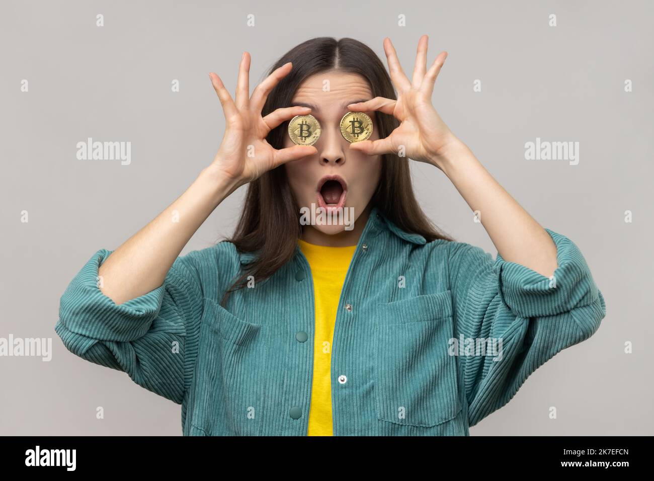 Porträt einer verblüfften, überraschten Frau, die die Augen mit goldenem Bitcoin bedeckt, die Kamera durch die BTC-Münze schaut, den Mund offen hält und eine Jacke im lässigen Stil trägt. Innenaufnahme des Studios isoliert auf grauem Hintergrund. Stockfoto