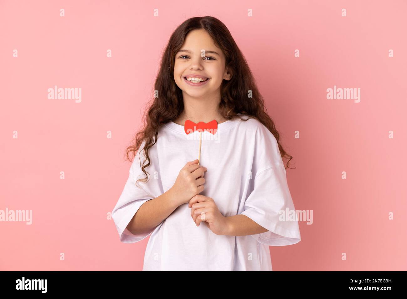 Porträt von zufriedenen kleinen Mädchen trägt weiße T-Shirt mit lustigen Papier Fliege, Spaß beim Spielen, trägt Maskerade Accessoire. Innenaufnahme des Studios isoliert auf rosa Hintergrund. Stockfoto
