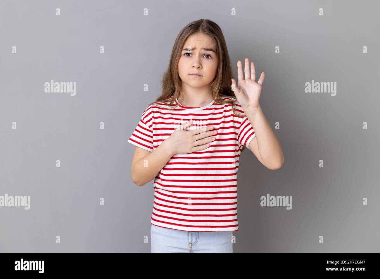 Porträt eines ernsthaften patriotischen kleinen Mädchens mit gestreiftem T-Shirt, das Hand am Herzen hält und die Wahrheit, Ehre und Gewissen sagt. Innenaufnahme des Studios isoliert auf grauem Hintergrund. Stockfoto