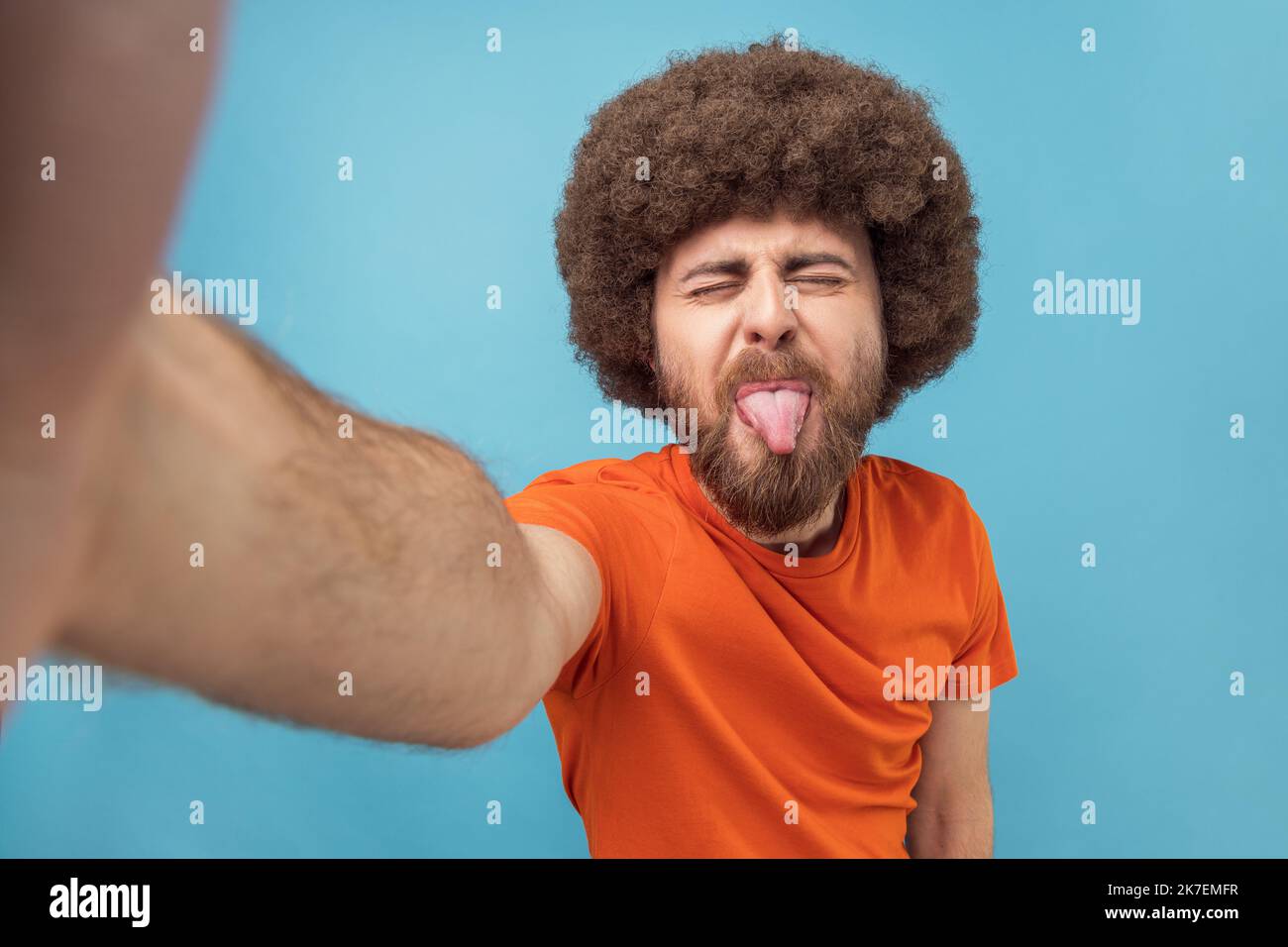 Porträt eines kindisch albernen Mannes mit Afro-Frisur in orangefarbenem T-Shirt, der Selfie macht oder Livestream macht, die Zunge zeigt, Sichtfoto. Innenaufnahme des Studios isoliert auf blauem Hintergrund. Stockfoto