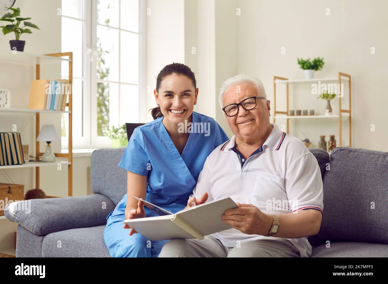 Glücklicher älterer Mann und junge Krankenschwester, die auf der Couch sitzen und gemeinsam Buch lesen Stockfoto