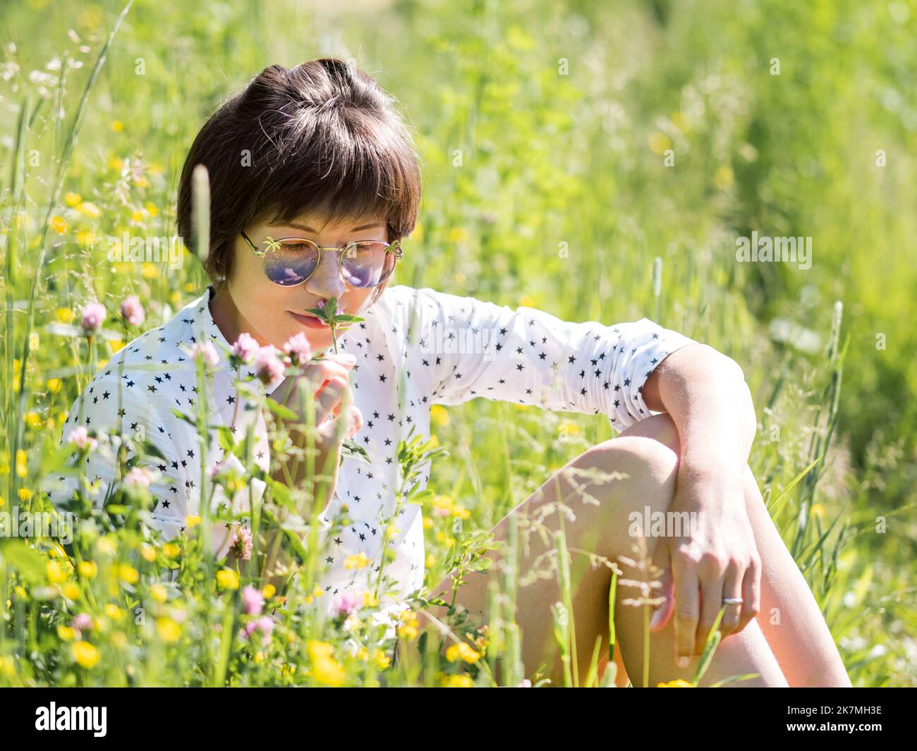 Frau in Sonnenbrillen, genießt Sonnenlicht und Blumenduft auf Grasfeld. Sommerliche Stimmung. Entspannen Sie sich im Freien. Selbstberuhigend. Stockfoto