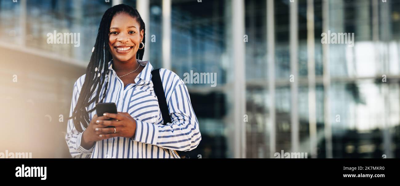 Glückliche junge Geschäftsfrau, die die Kamera anlächelt, während sie zur Arbeit in der Stadt pendelt. Porträt einer fröhlichen jungen Geschäftsfrau mit Smartphone auf Stockfoto