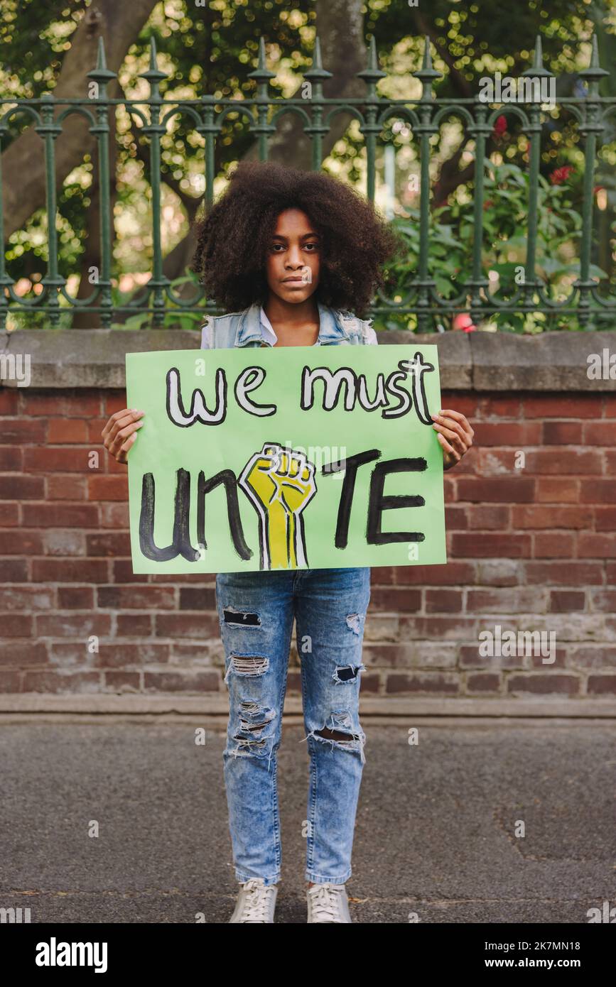 Junges schwarzes Mädchen, das die Kamera anschaut, während es ein Plakat hält, das zur Einheit aufruft. Jugendaktivistin protestiert gegen Rassenungleichheit und Diskriminierung Stockfoto