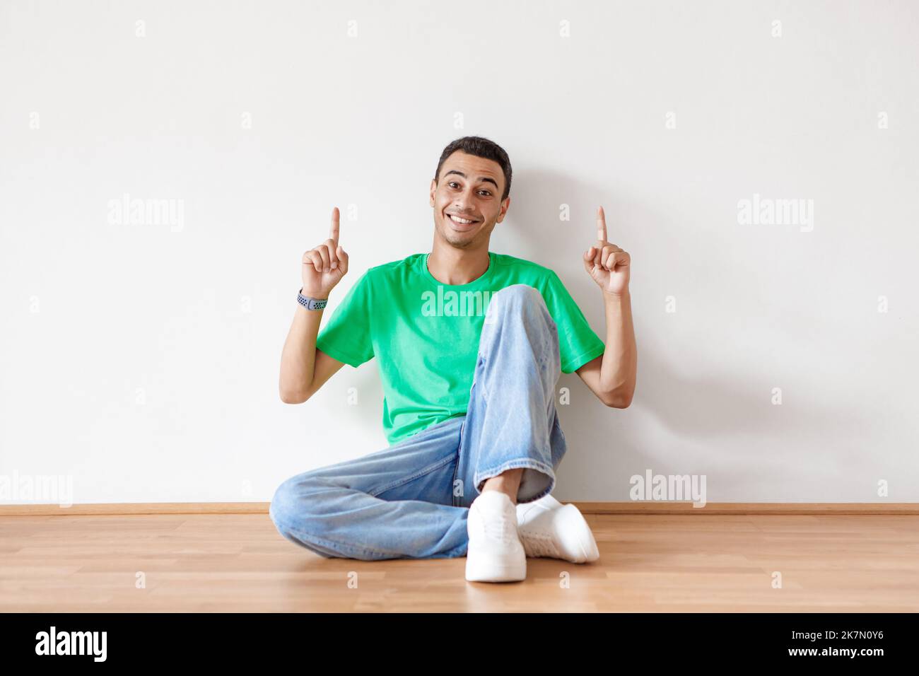 Fröhlicher arabischer Mann, der auf dem Boden sitzt und auf den freien Platz über ihm zeigt, um Werbung zu machen, und lächelt über der weißen Wand auf die Kamera Stockfoto