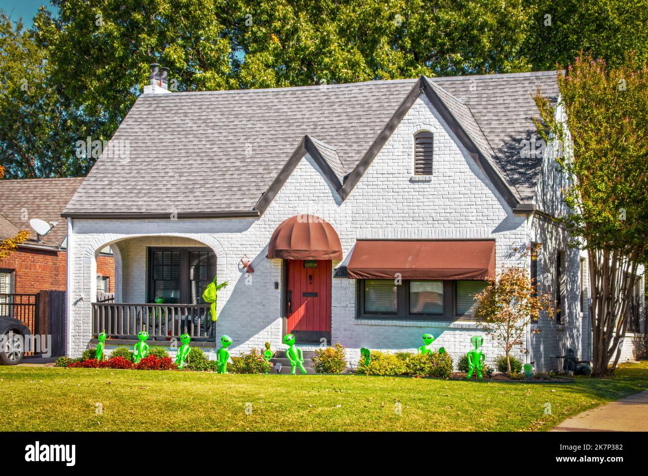 Niedliches Häuschen aus weiß lackiertem Ziegel mit Markisen und roter rustikaler Tür, die für Halloween mit aufblasbaren grünen Außerirdischen dekoriert ist - Invasion aus dem Weltraum Stockfoto