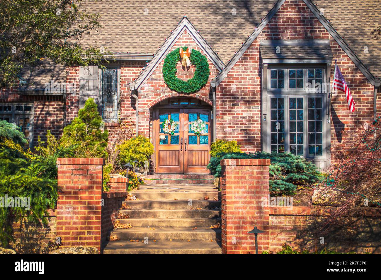 Rustikales gehobenes Ziegelwohnhaus mit Magnolienblütenkranz an Doppeltüren und großem Weihnachtskranz oben - mit amerikanischer Flagge - Nahaufnahme Stockfoto