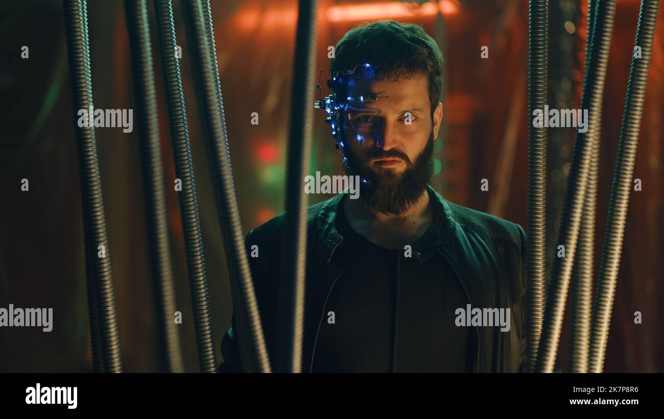 Junge Brünette Kerl im Cyberpunk-Stil steht hinter den schwarzen Stäben hängen. Ein futuristisches Headset mit Okular und Mikrofon tragen. Neonlicht Hintergrund. Science-Fiction. Stockfoto